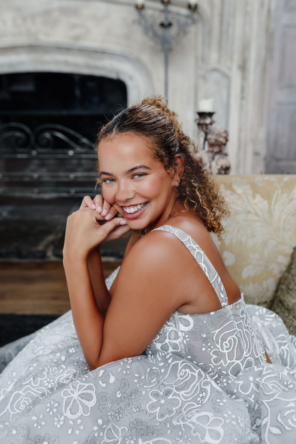 A bride wearing a white floral lace wedding dress poses on stone steps with natural lighting.