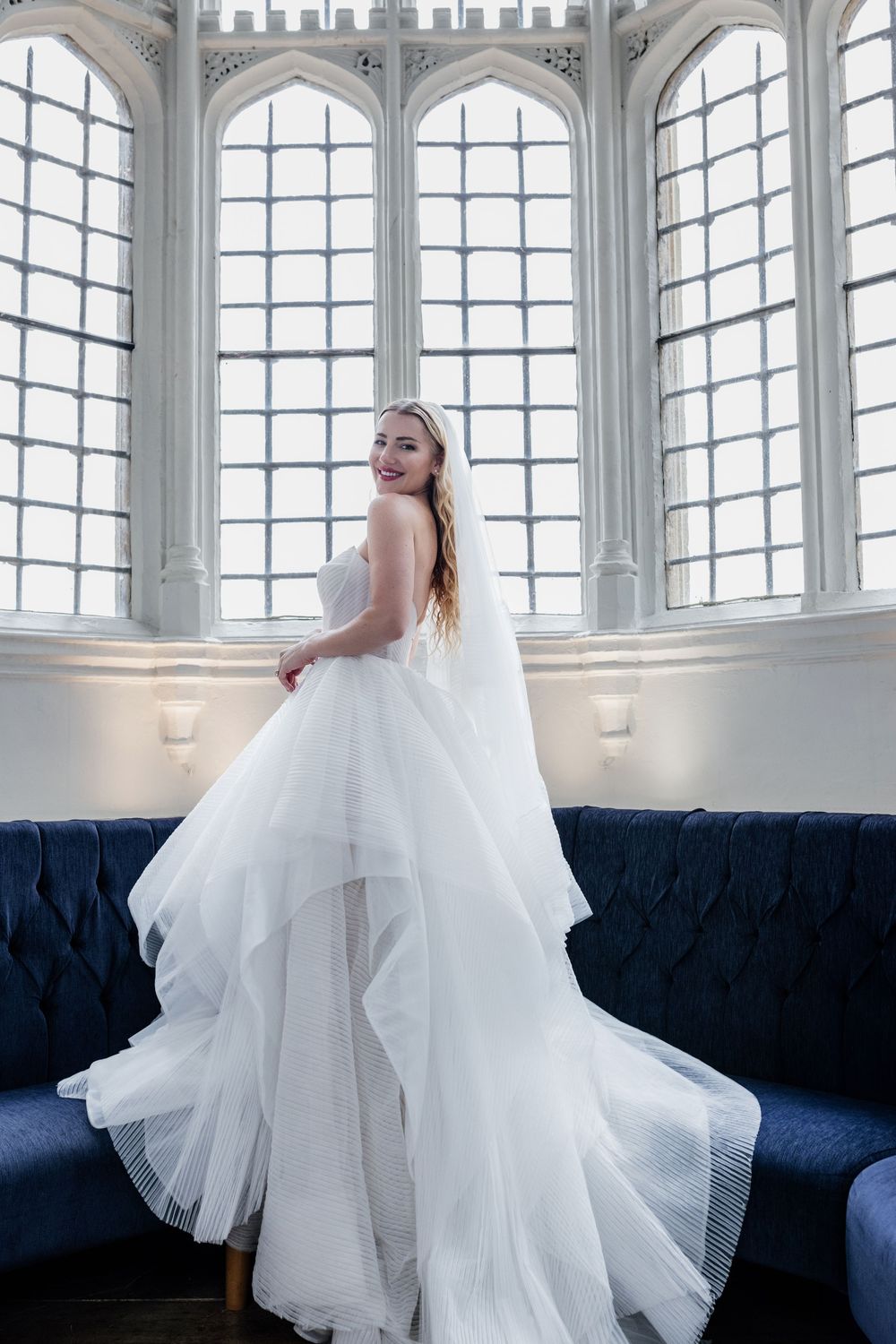 A flowing white wedding dress with voluminous tulle layers is displayed in an elegant room with arched windows.