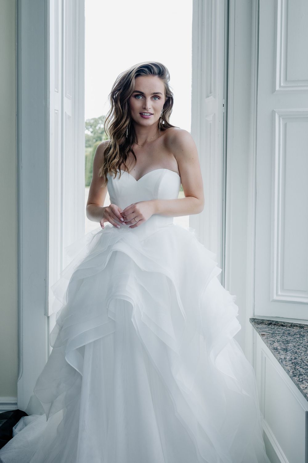 Elegant bride in voluminous white wedding gown poses in bright sunlit room with architectural details.