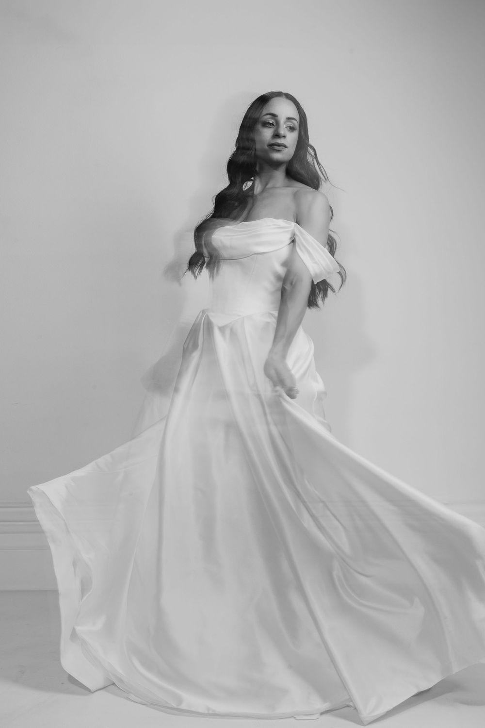 A graceful figure twirls in an elegant off-shoulder white wedding gown with flowing fabric in a black and white portrait.
