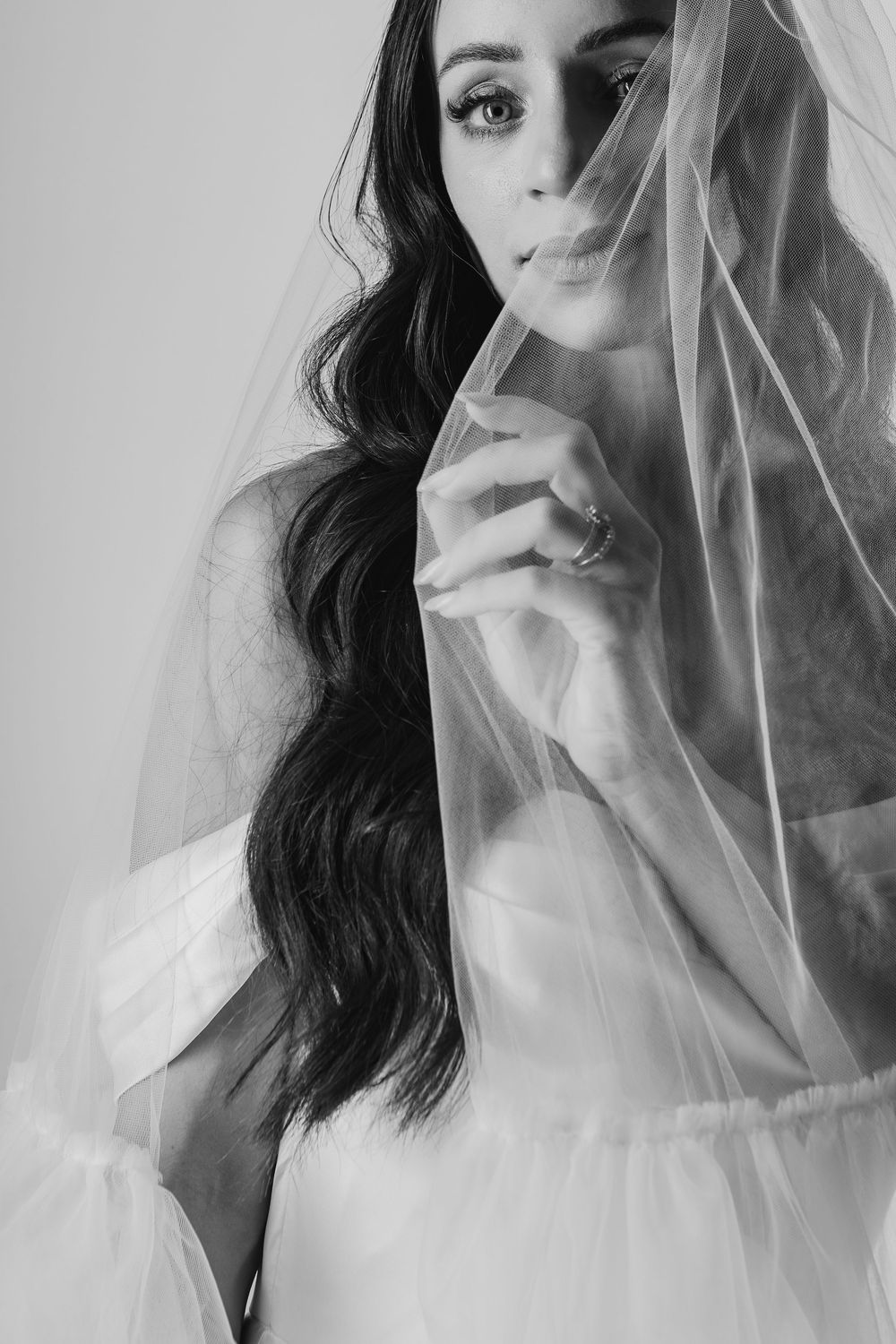 Ethereal black and white bridal portrait with flowing veil and long dark wavy hair against light background.