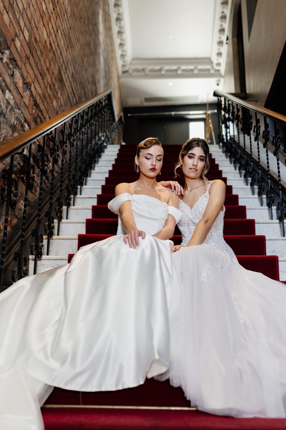 Two people in white wedding dresses sit on elegant staircase with red carpet and ornate black railings.