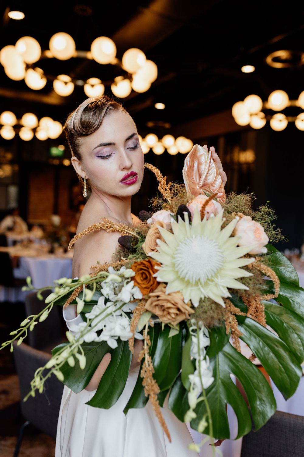 Elegant floral arrangement featuring white spider mums, monstera leaves and golden accents in moody lighting.