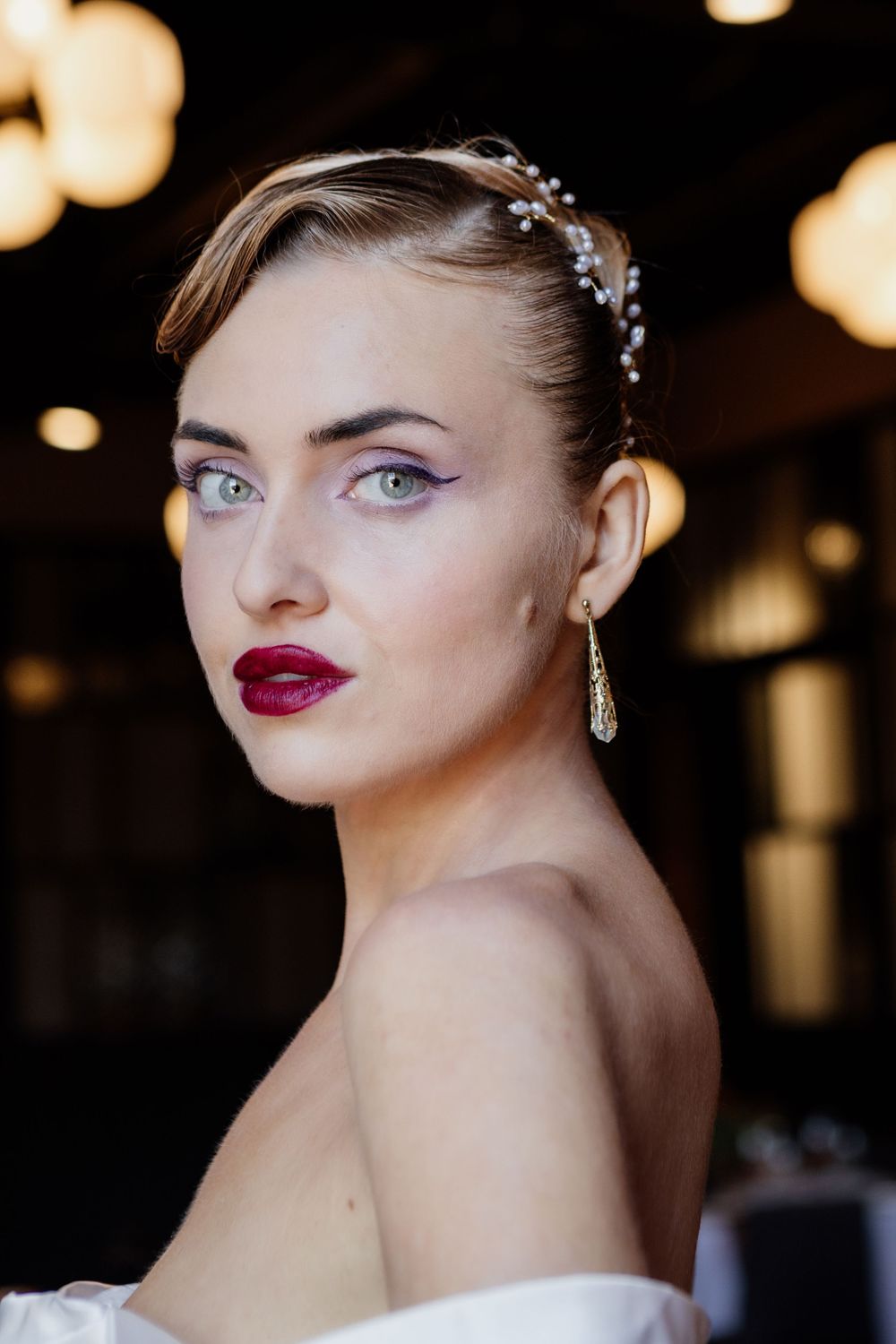 Elegant bridal portrait with sparkly hairpiece, red lipstick and dangling earrings against blurred bokeh lights.