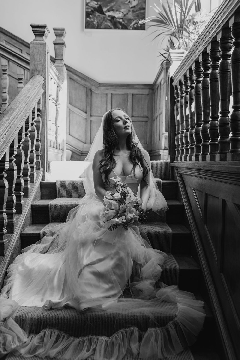 Dramatic black and white photo of a bride sitting on vintage stairs in a flowing tulle gown holding a bouquet.