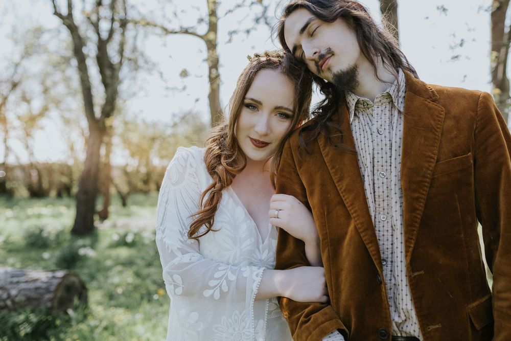 A couple in fall attire embraces outdoors with a lace wedding dress and brown corduroy jacket against autumn foliage.