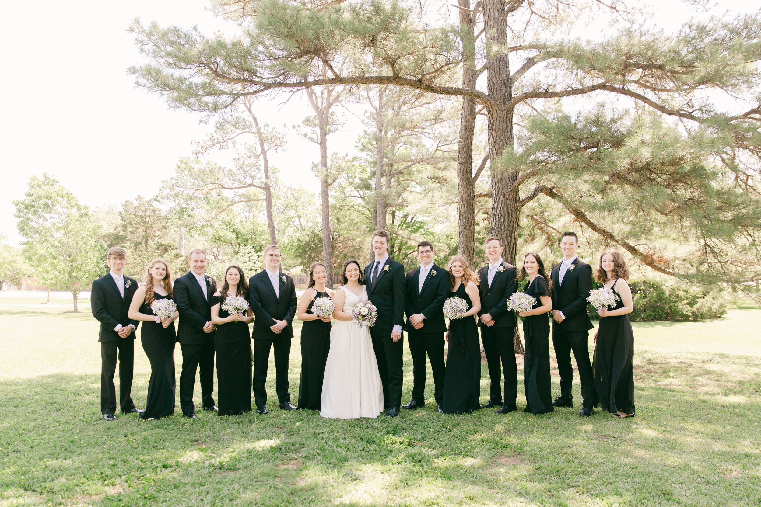 Wedding party in black attire lined up outdoors under towering pine trees.