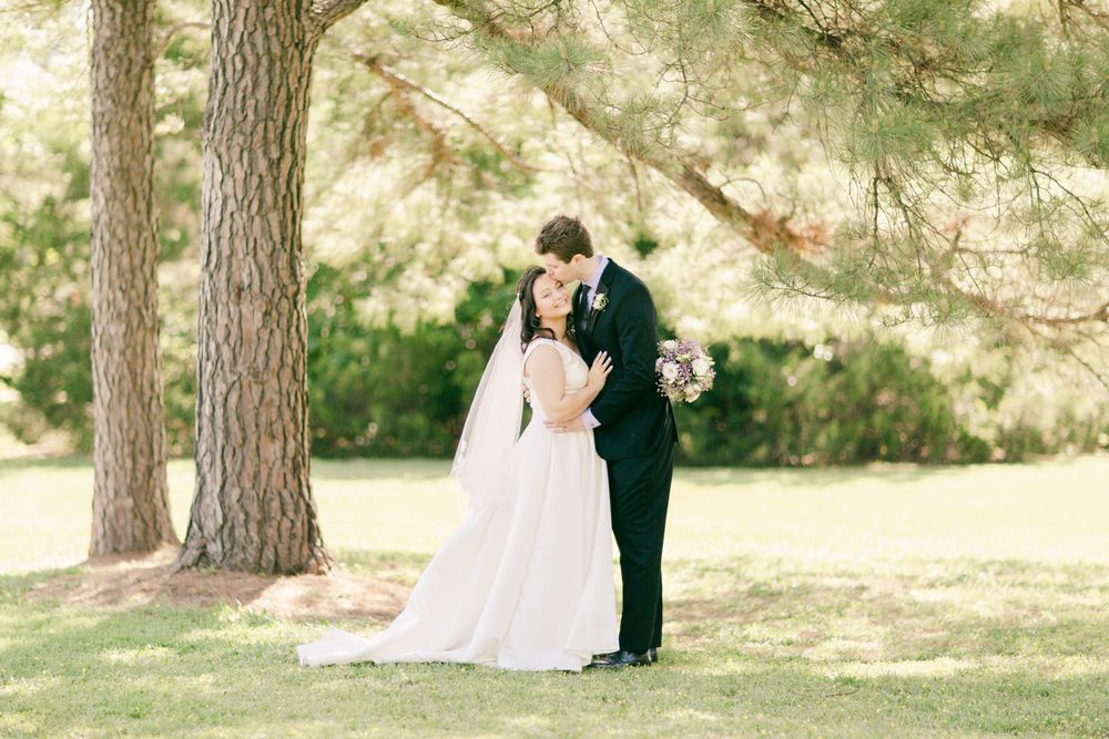Romantic wedding couple embracing under tall pine trees in a sunlit garden setting.