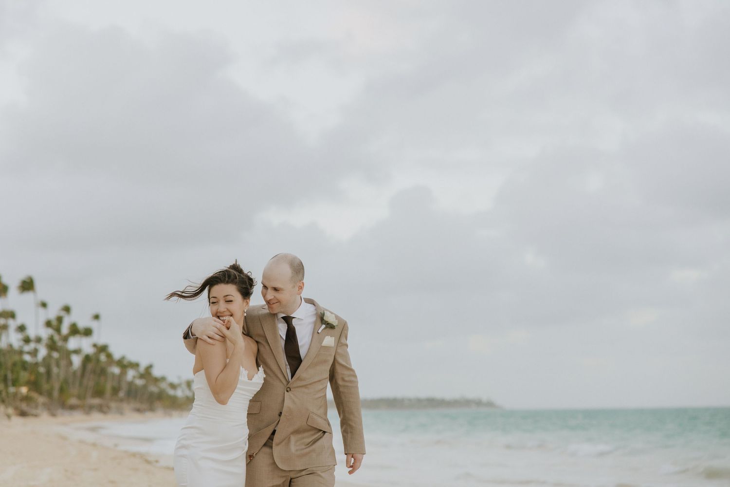 A couple in light beige attire walks together along a scenic tropical beach with palm trees and ocean waves.