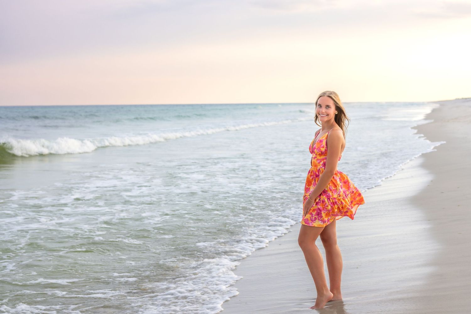 A person stands at the edge of the ocean wearing a summery orange dress as waves roll in during sunset.