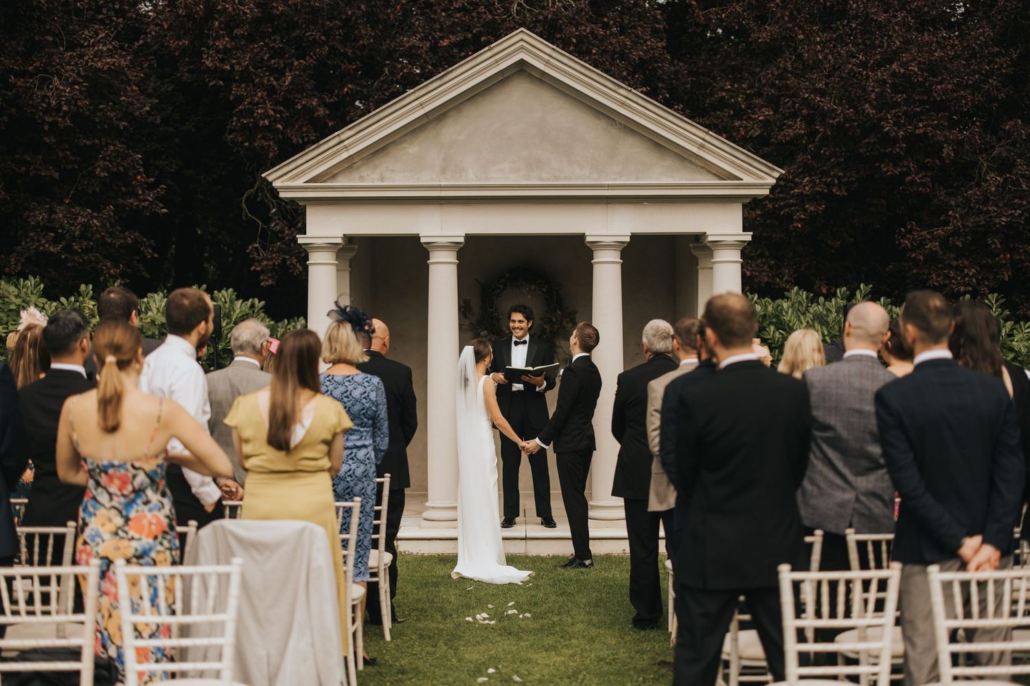 Outdoor wedding ceremony with a white columned gazebo and rows of white chairs set up on a grassy lawn.