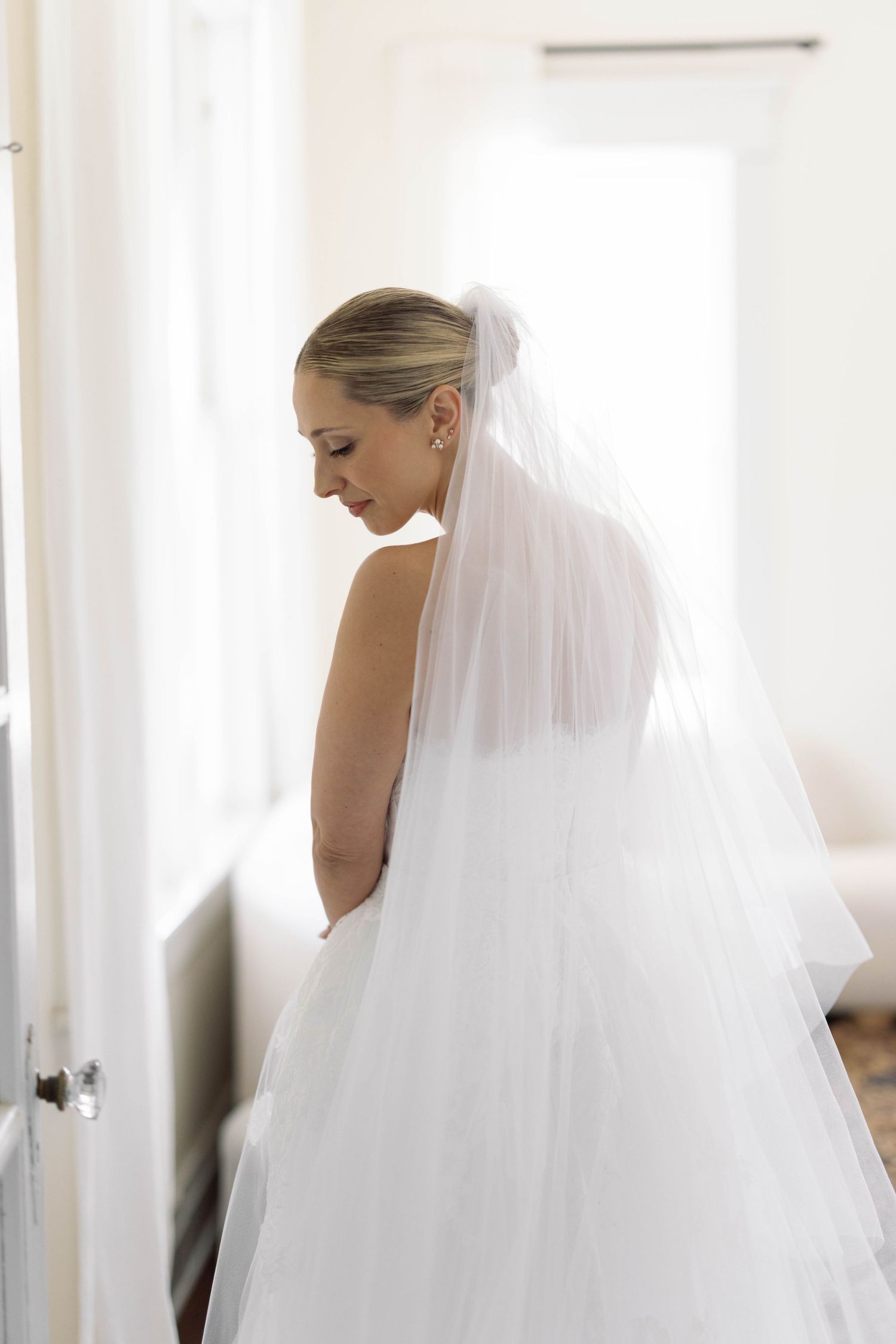 A bride in a white wedding dress and flowing veil stands in profile against a bright window on her special day.