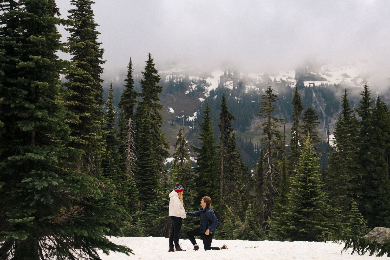 Two people stand in snowy mountain landscape surrounded by tall evergreen trees and snow-capped peaks in the background.