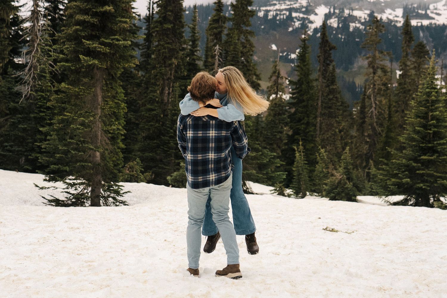 A couple embraces in a snow-covered forest with evergreen trees and mountain views in the background.