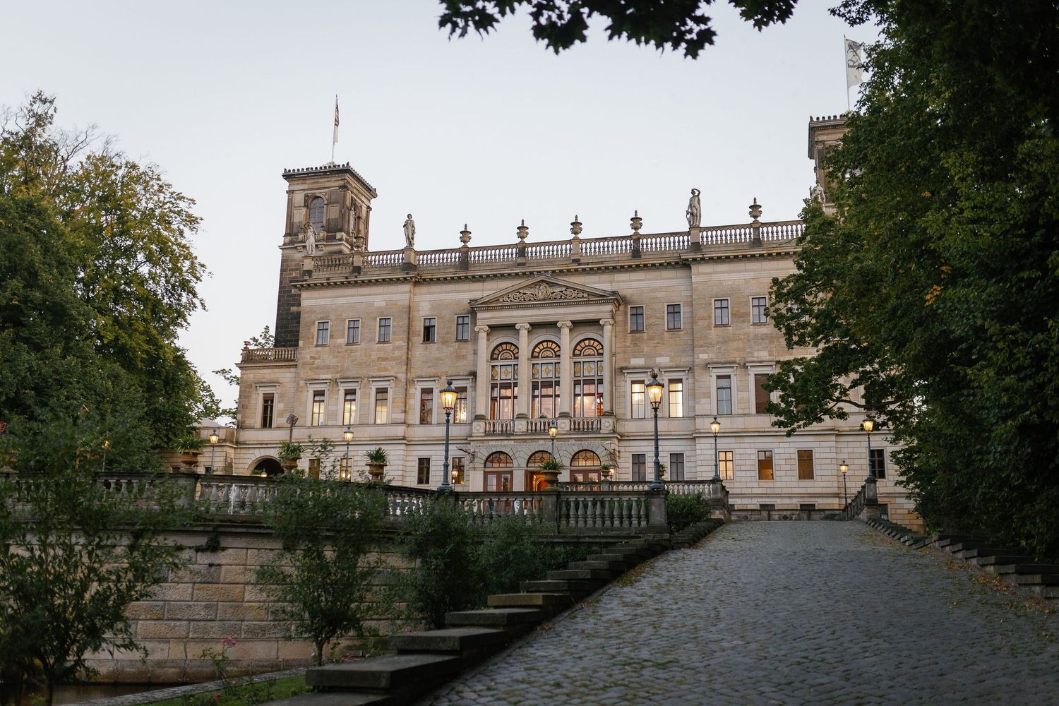 An ornate medieval castle exterior with stone walls and terraced gardens shown in a panoramic view during daytime.
