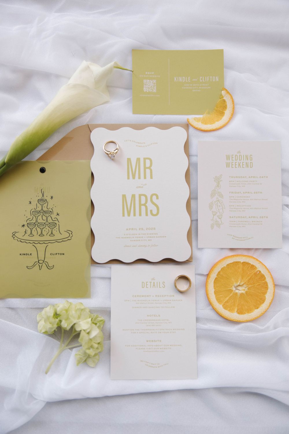 Elegant wedding stationery suite in soft sage green and cream with orange slices and floral accents on light fabric.