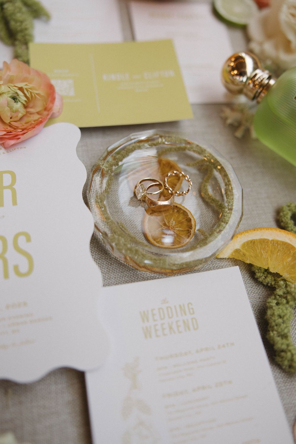 Wedding bands and engagement rings rest delicately on white and gold wedding stationery with fresh blooms and moss accents.