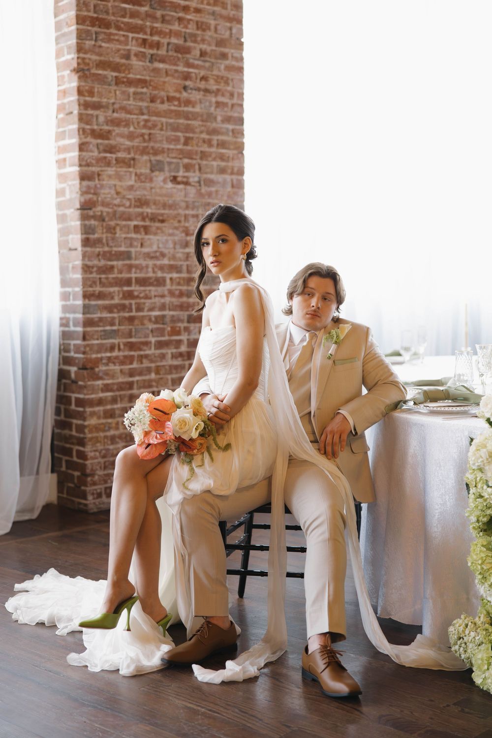 Modern styled wedding couple in white outfits sit atop industrial brick loft table with a peach floral bouquet.