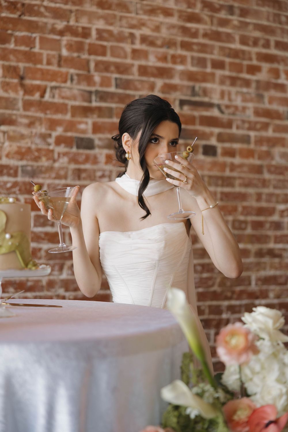 A person in a white gown holds up a wine glass against a rustic brick wall at an elegant event.