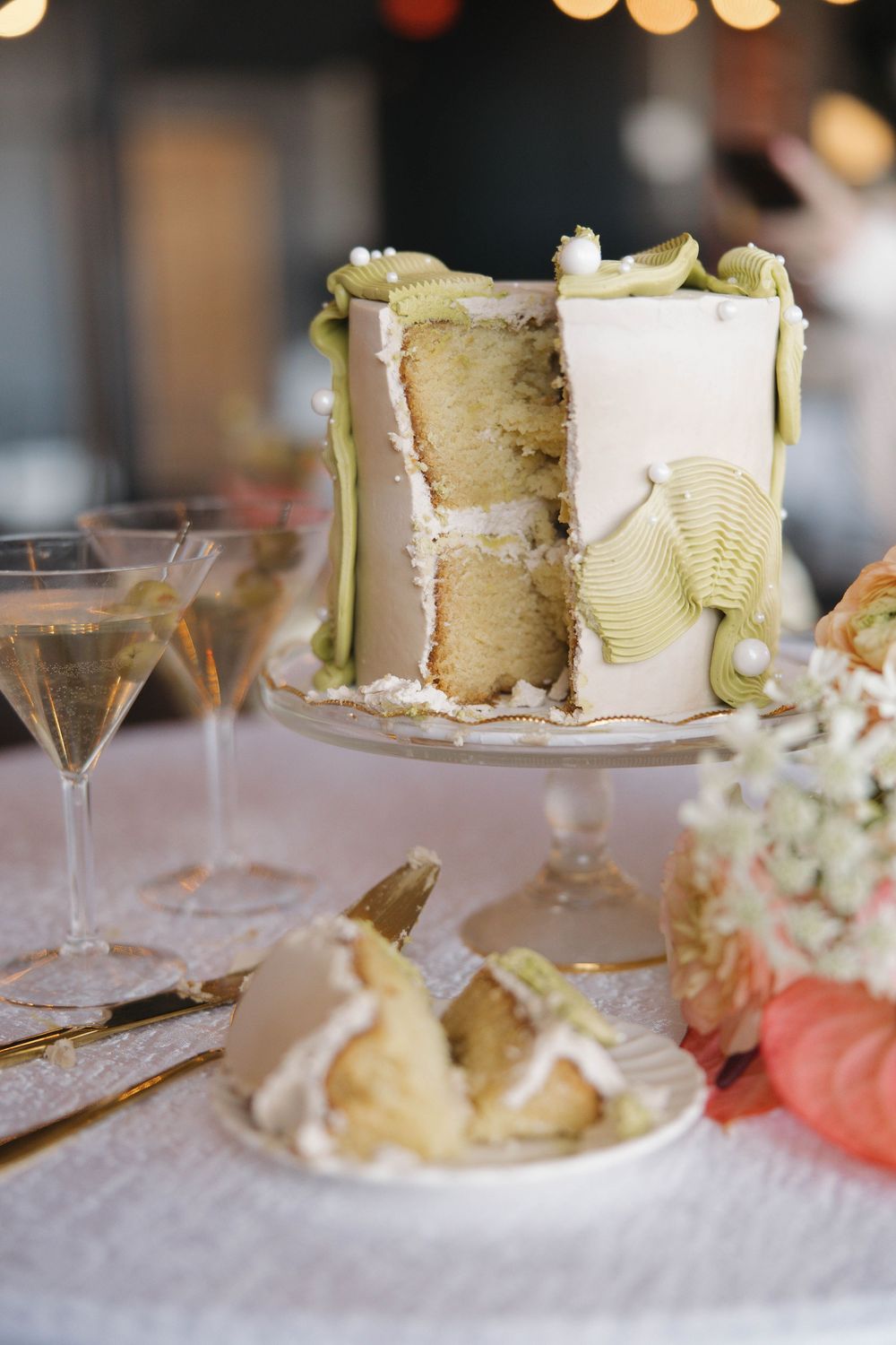 Elegant white wedding cake decorated with green ribbons and accents displayed on a glass pedestal next to champagne glasses.