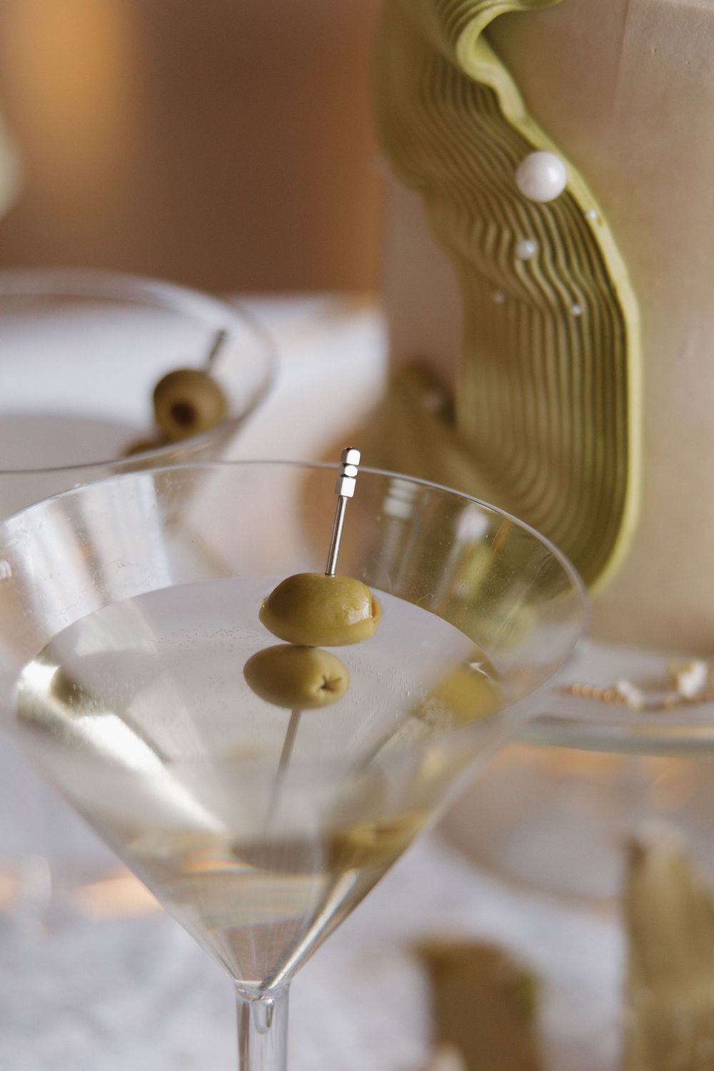 Olive garnish being added to a martini glass filled with clear cocktail and secured with a decorative toothpick.