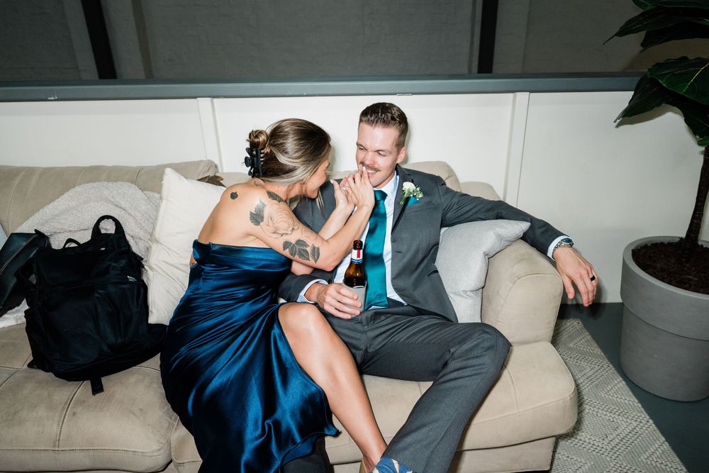A couple shares a playful moment on a beige couch, the woman wearing a navy blue evening gown.