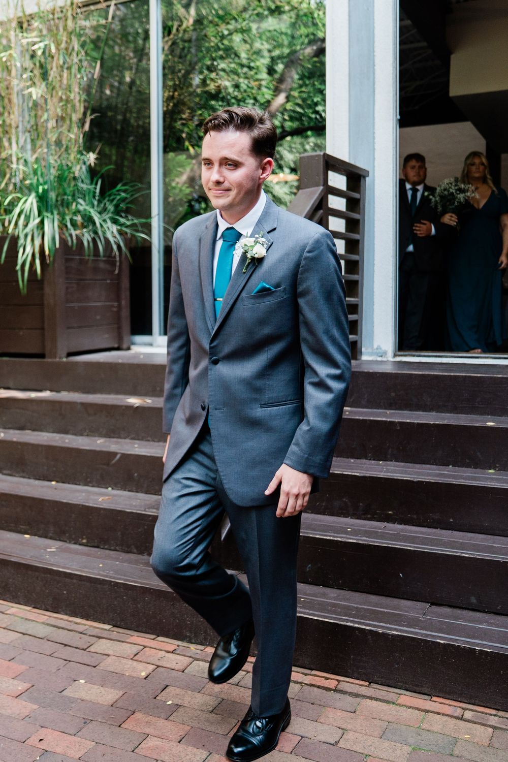 Groom in dark suit performs playful dance moves on brick steps outside building with windows.