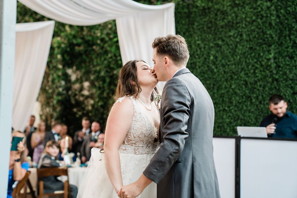 A romantic wedding ceremony moment under a white draped arch against a green hedge wall with guests seated in the background.