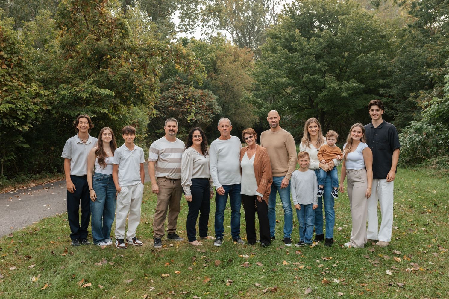 Large family group portrait outdoors in autumn park setting, posing together on green grass with fall foliage trees.