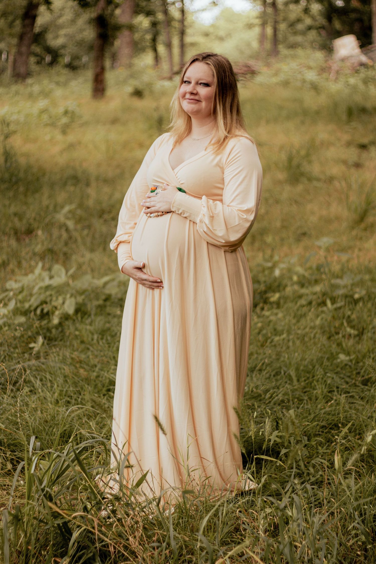 An expectant mother in a flowing cream-colored dress stands thoughtfully in a sunlit wooded meadow.