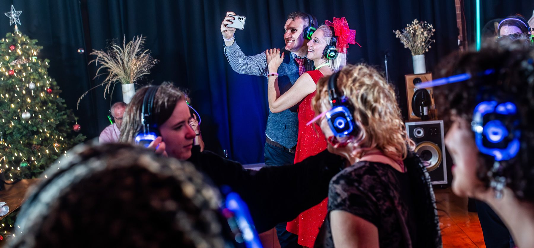 People celebrate at a holiday party with a decorated Christmas tree and blue lights in a dark room.
