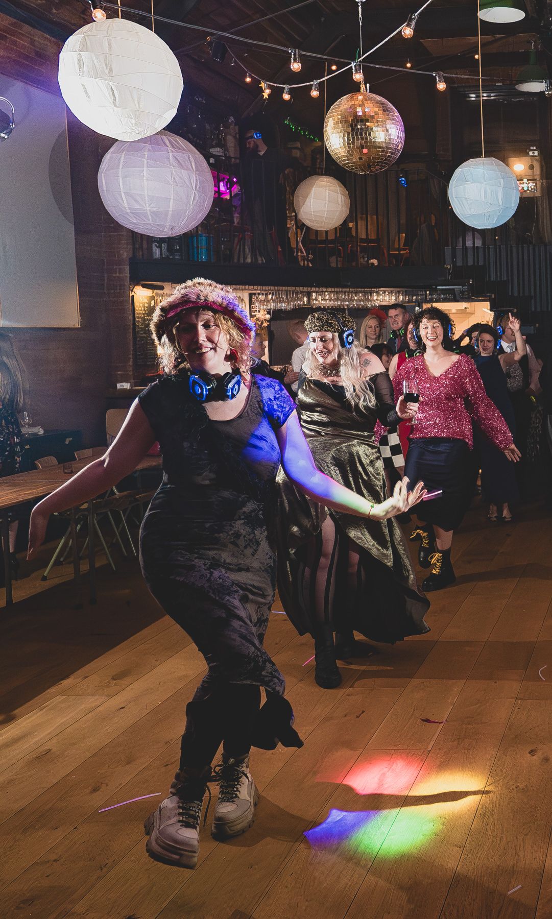 Dancers move energetically on a wooden dance floor under white paper lanterns and a disco ball with colorful lighting.