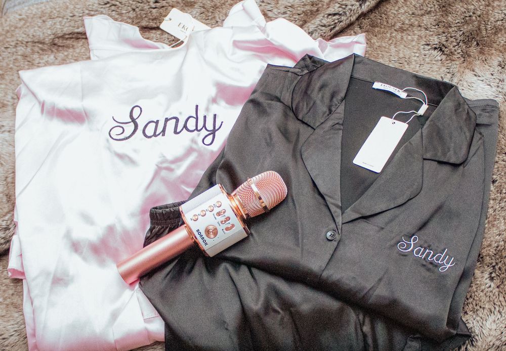 White and charcoal silk robes with embroidered names and a matching pink water bottle displayed on textured surface.