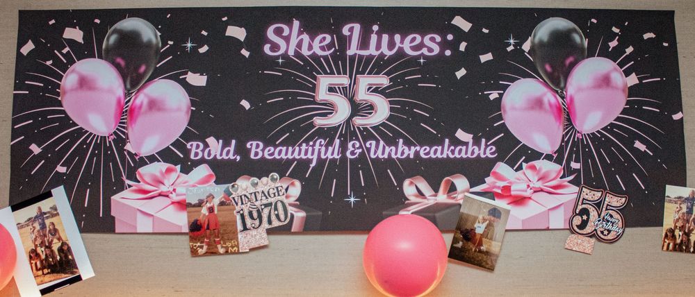 A decorative banner with pink and black balloons featuring the text 'She Lives' and numbers against a dark background.