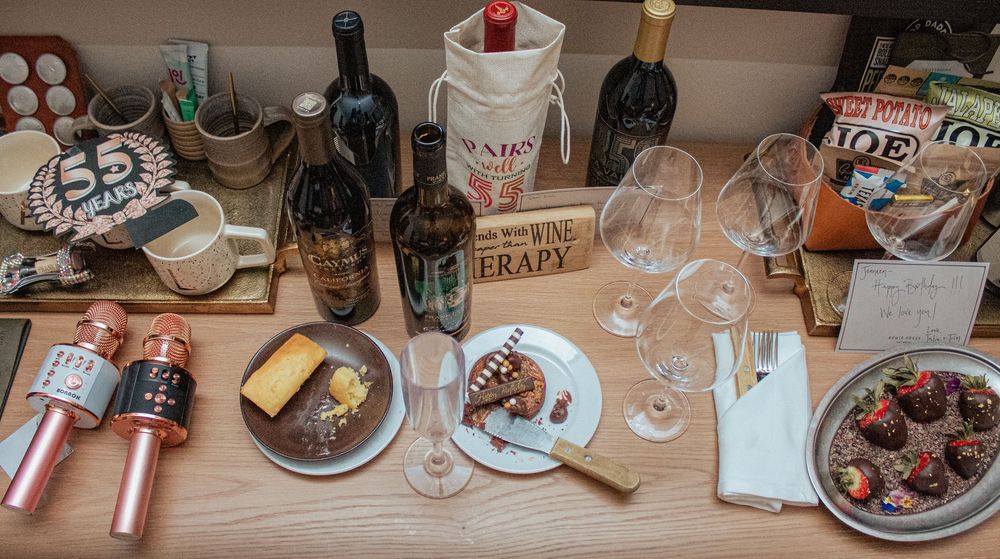 A wooden table set with wine bottles, glasses, cheese plates and appetizers for a gathering.