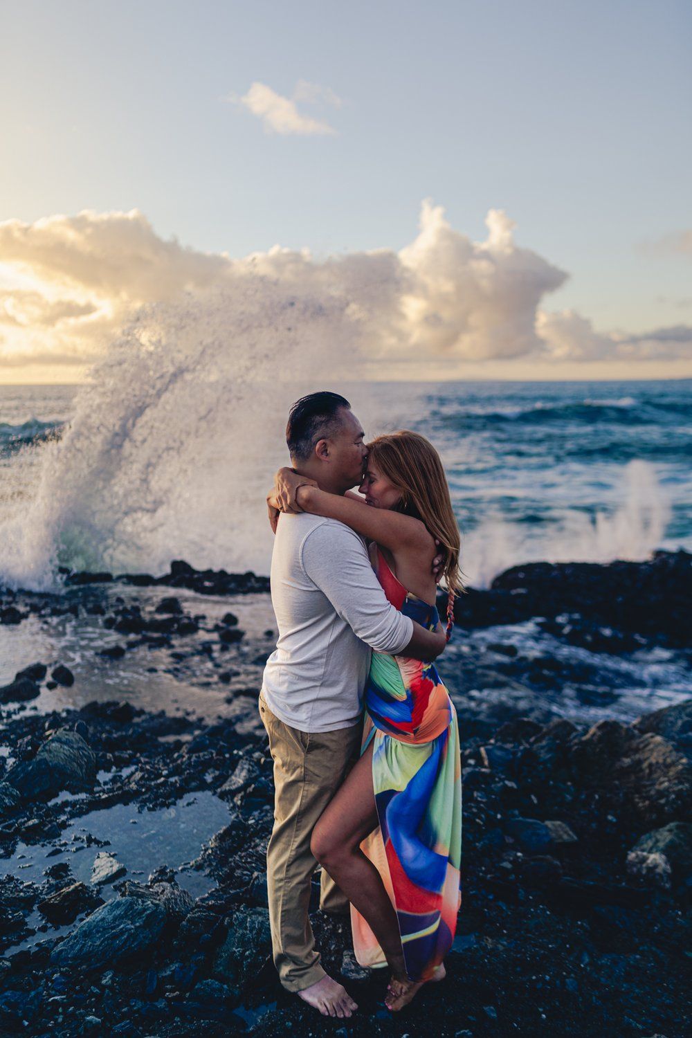 A romantic couple embraces on rocky shore at table rock laguna beach sunset as ocean waves crash  and spray in the background.