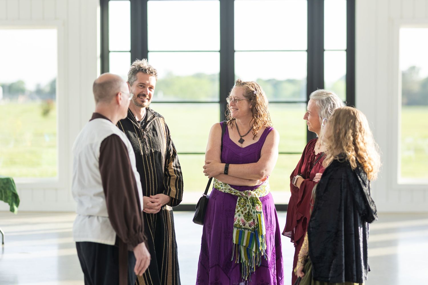 A small group of people in formal attire gather in conversation near large windows in a bright interior space.
