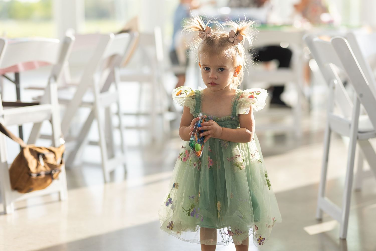 A small child wearing a mint green tulle dress with fluttering details stands in a sunlit room with white folding chairs.