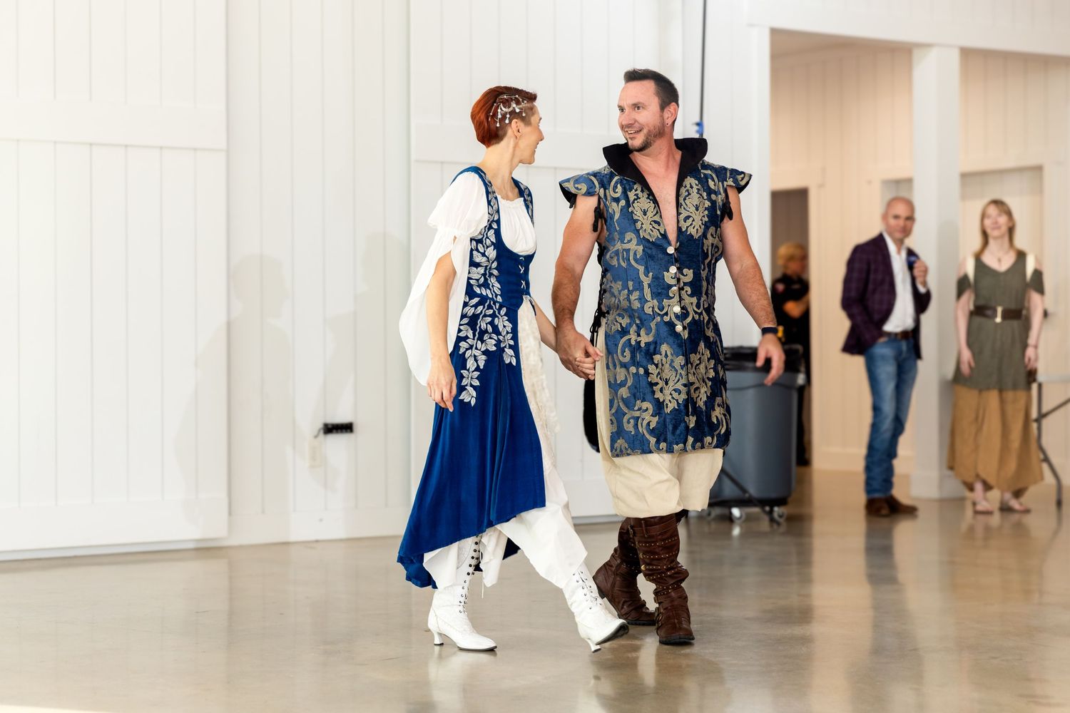 Two dancers in coordinating blue and white Renaissance-style costumes perform together in a bright indoor space.