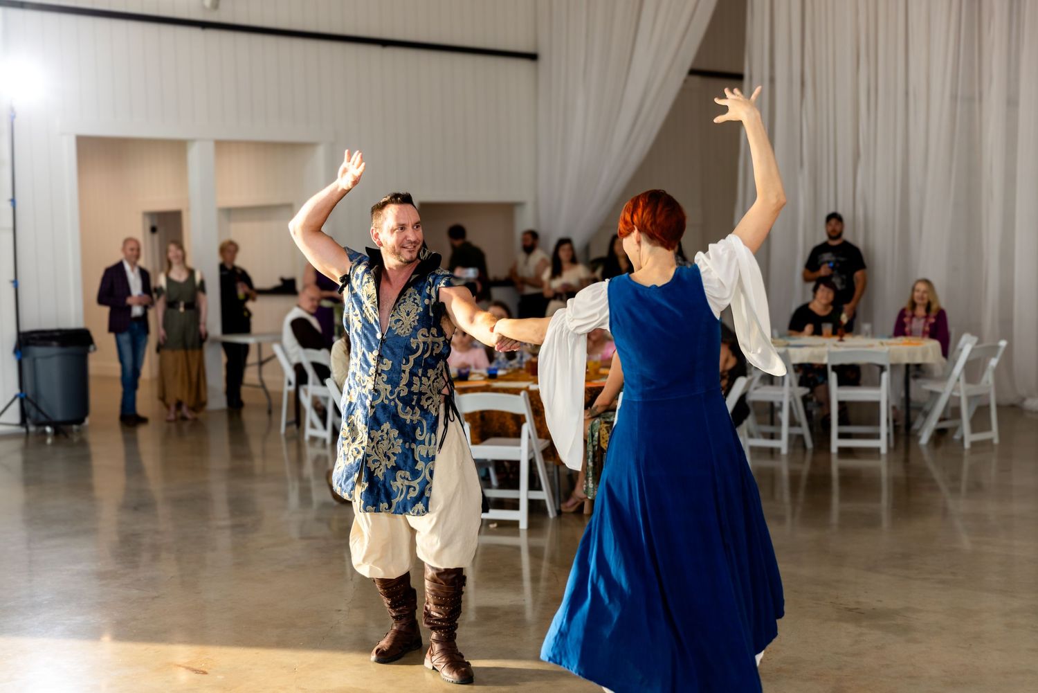 Two people enthusiastically dancing together at a reception venue with white chairs and curtains in the background.