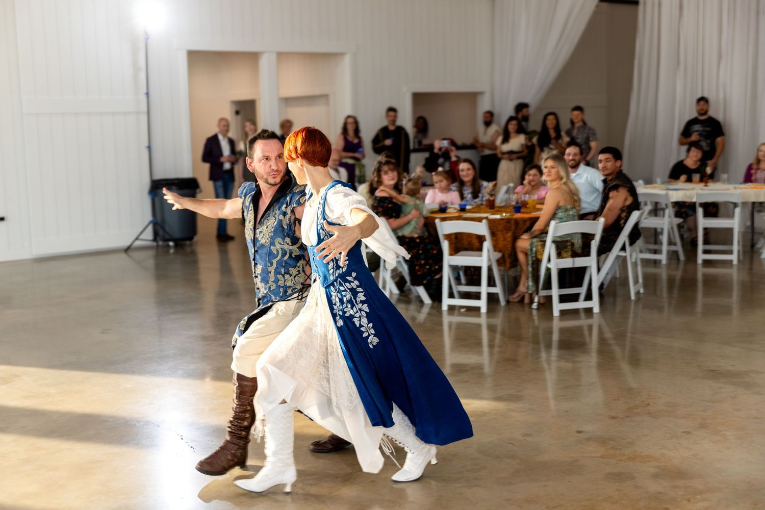 Dancers in a ballroom perform choreographed moves while guests watch from white chairs in an elegant venue.