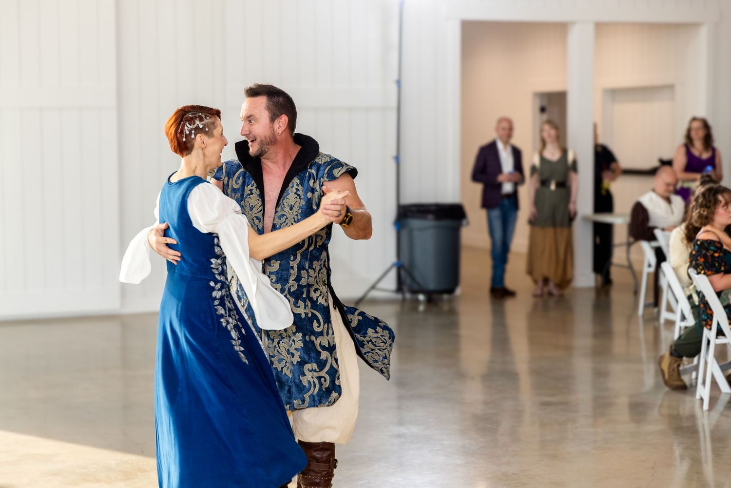 Dancers in matching medieval-inspired blue and white outfits performing a traditional dance in a bright hall.