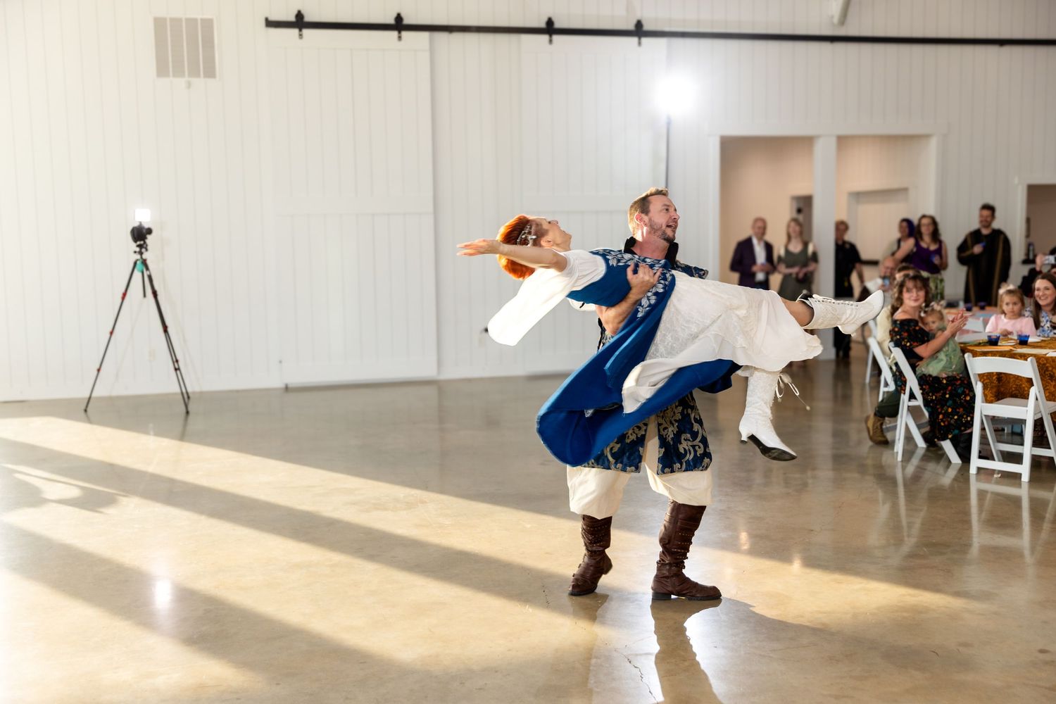 A group performs modern dance movements in a bright studio space while onlookers watch from white chairs along the wall.