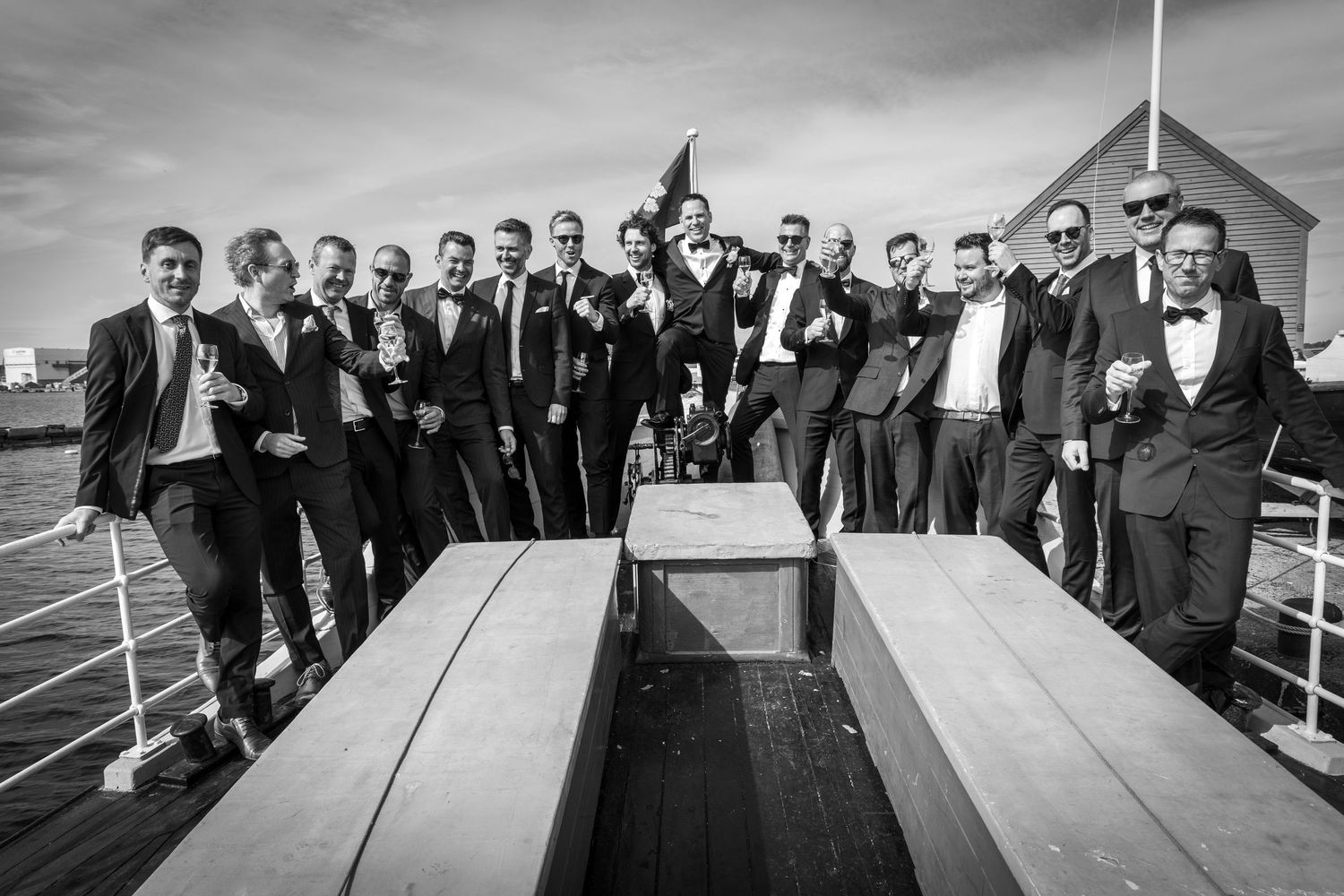 Large group of groomsmen in tuxedos celebrate on waterfront pier, raising glasses in festive toast.