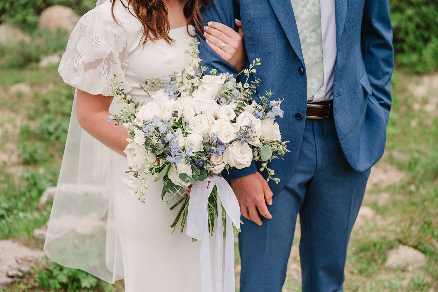 A stunning white bridal bouquet with roses and delicate greenery held against a navy blue suit and cream dress.