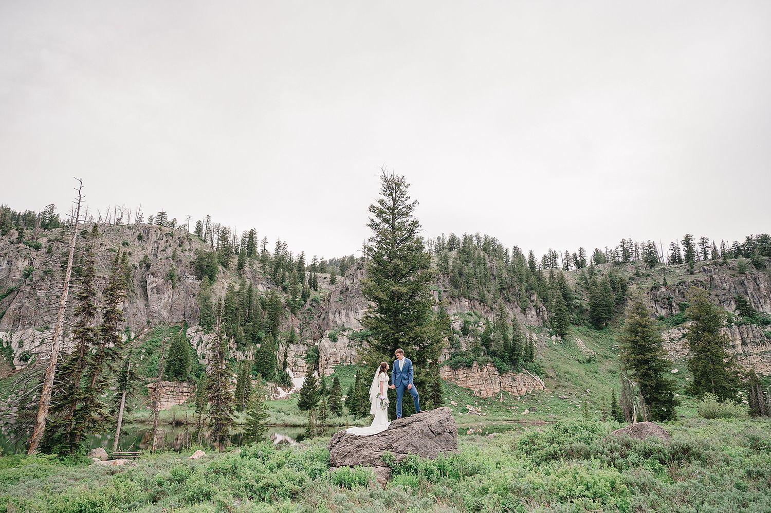 A couple stands in a lush alpine meadow surrounded by evergreen trees and rocky mountain peaks.