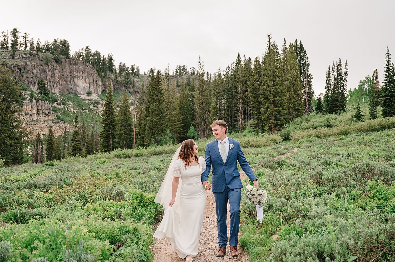 Bride and groom taking romantic wedding photos in a mountain meadow with towering peaks in the background.