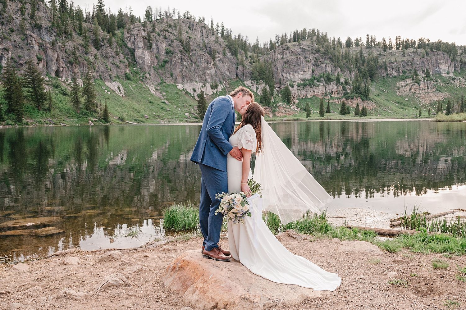 A couple stands together by an alpine lake surrounded by evergreen forests and mountain peaks in a scenic mountain valley.