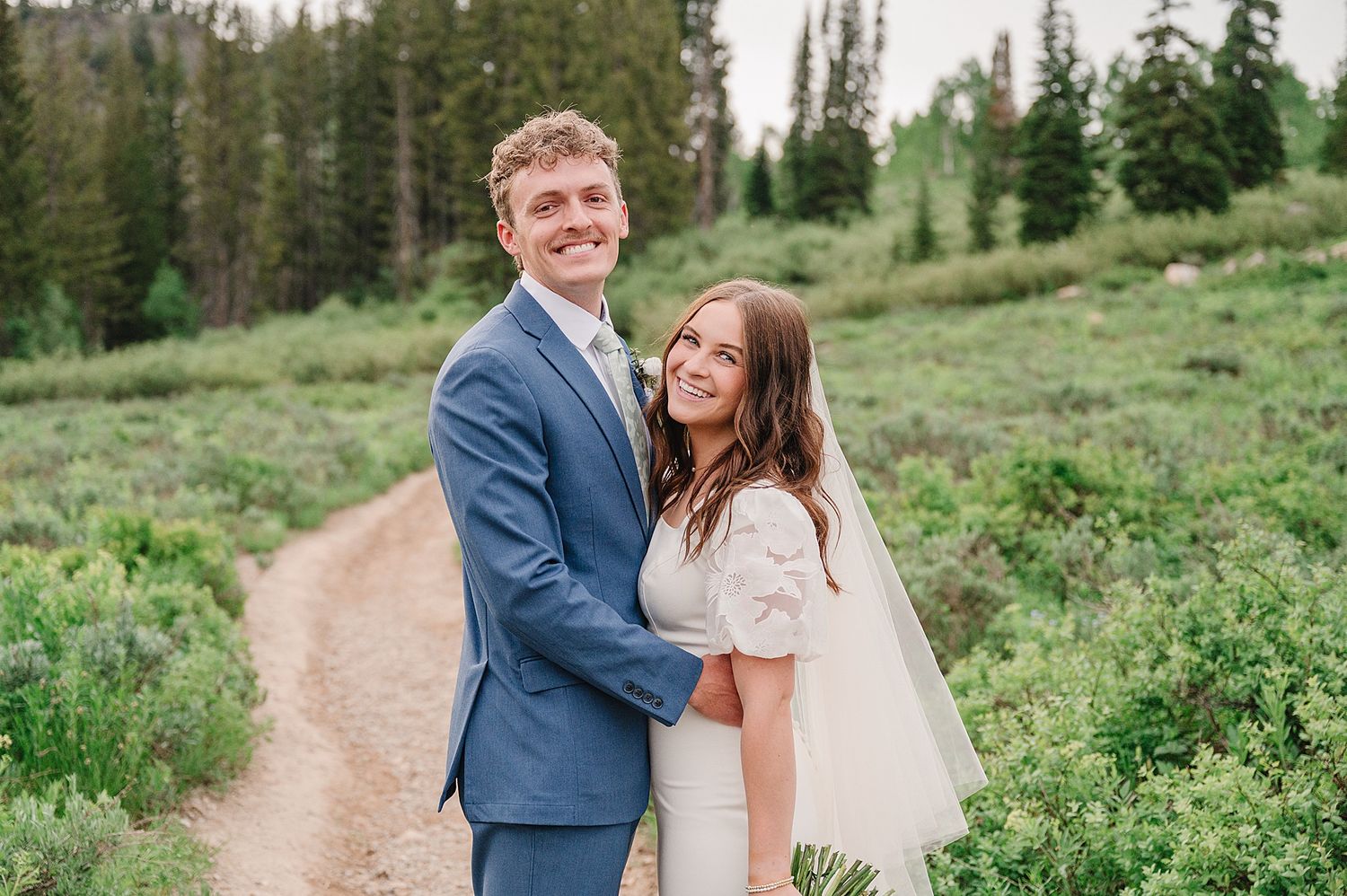 Newlyweds stroll along a scenic mountain path surrounded by evergreen trees and wildflowers on their wedding day.