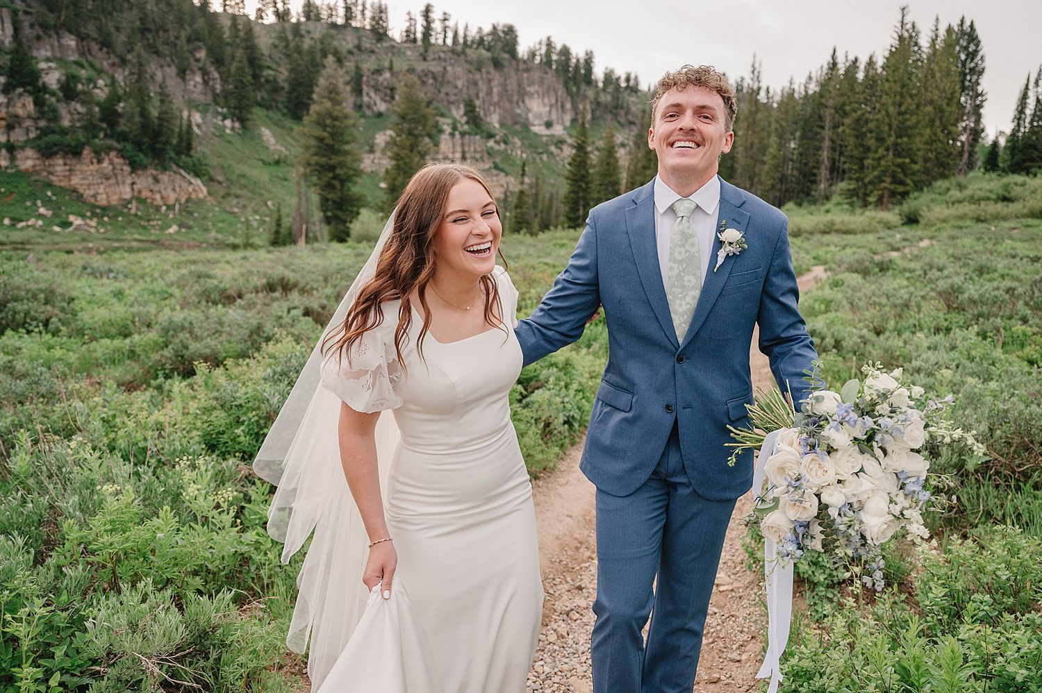 A bride and groom walk together on a mountain path surrounded by wildflowers and evergreen trees in a scenic setting.